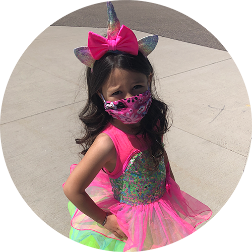 Child in a pink unicorn costume with face mask, standing outdoors on a sunlit pavement.