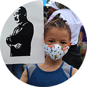 Child wearing a patterned face mask at a rally, with a black silhouette sign in the background