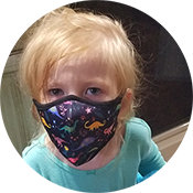 Blond child wearing a colorful face mask and teal shirt, looking at the camera indoors