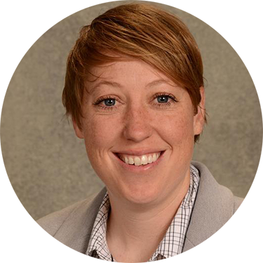 Smiling person with short light-brown hair in a gray blazer and checkered shirt, against a neutral background