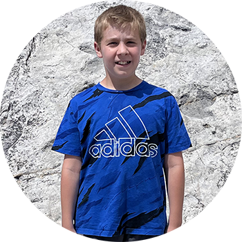 Boy in a blue Adidas shirt standing against a light stone wall.