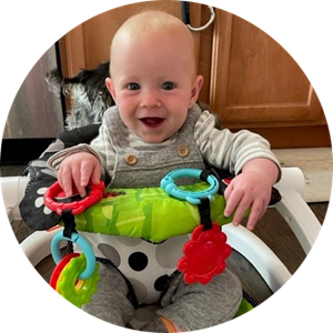 Smiling baby in a high chair, wearing gray overalls and holding colorful toys