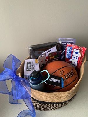 Basket of sports snacks and gear with a basketball, water bottle, snacks, and blue ribbon on a table