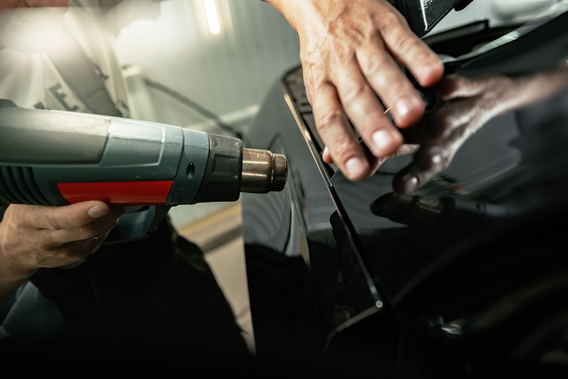 Hands using a heat gun to wrap black vinyl on a car panel