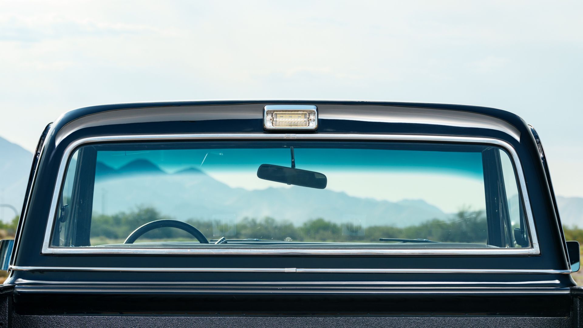 Rear window of a vintage blue car, showing a rearview mirror and distant mountains through the glass.