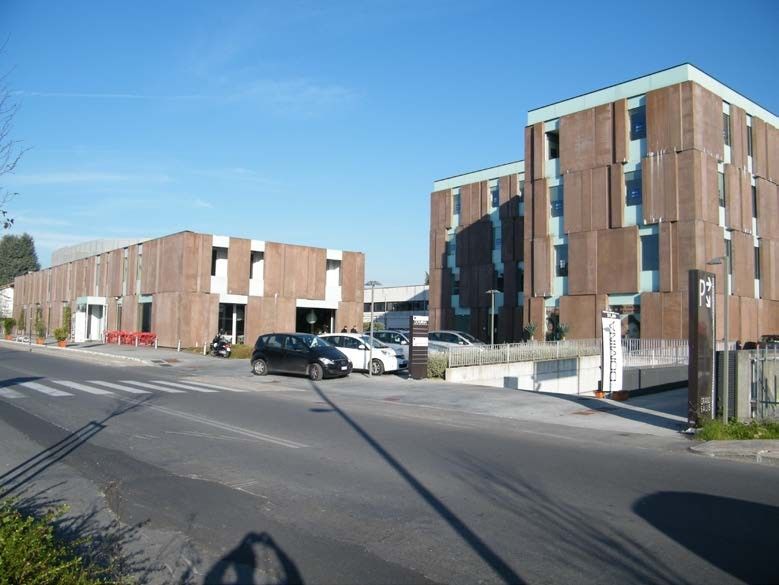 Due moderni edifici marroni con auto parcheggiate di fronte, visti da una strada con attraversamento pedonale.