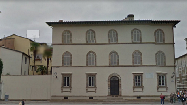 Edificio color crema a tre piani con finestre ad arco, porta scura e tetto piano in un contesto urbano.