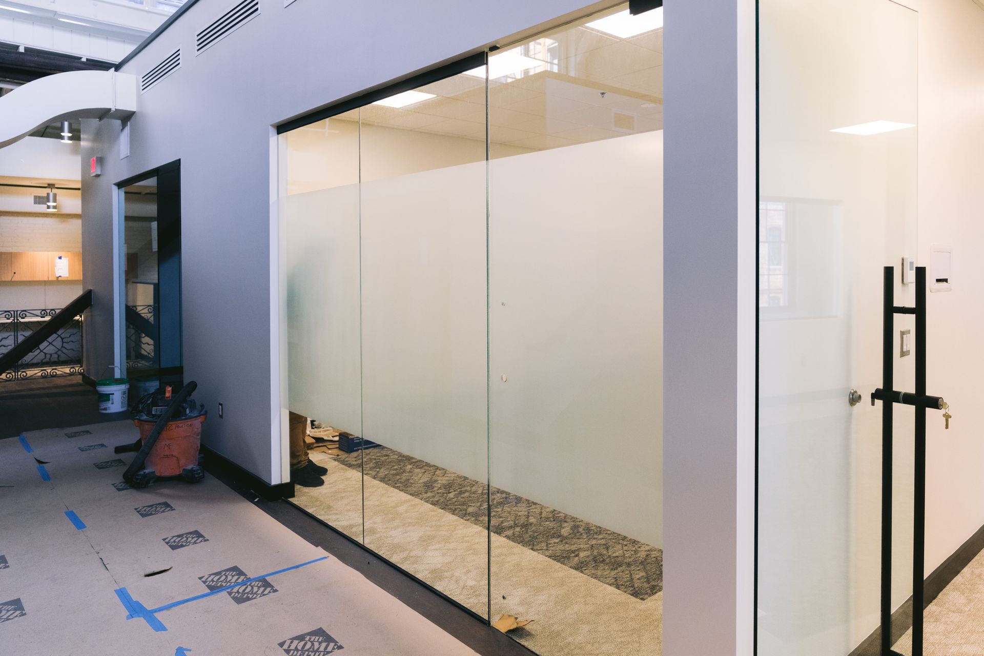 Glass-walled office corridor with white walls, a black door handle, and protective floor covering under construction
