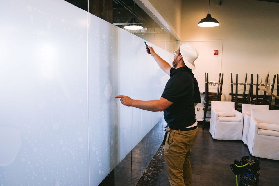 Person painting a large white wall in a bright room with stacked chairs and supplies nearby