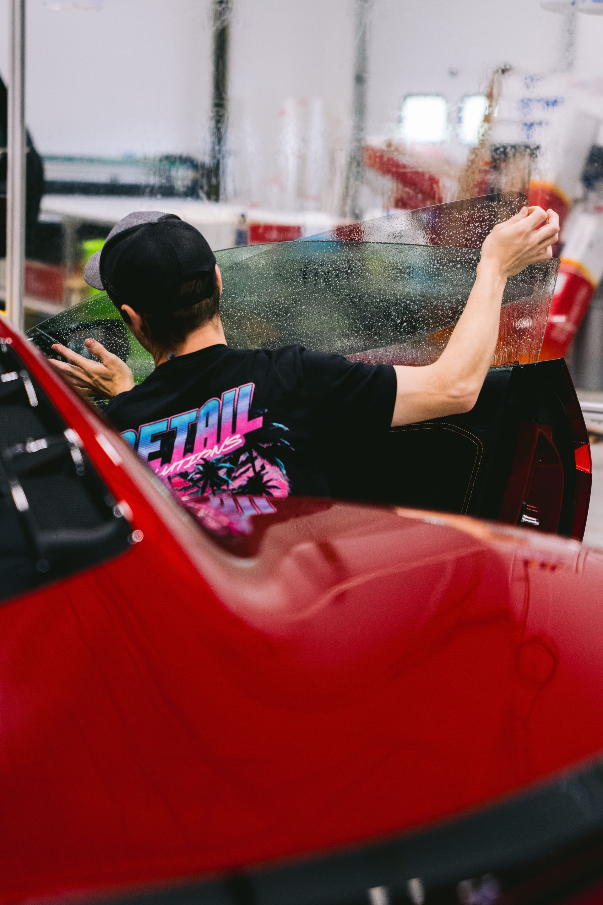 Person in black T-shirt leans out of a red car, reaching toward a colorful spray of water.