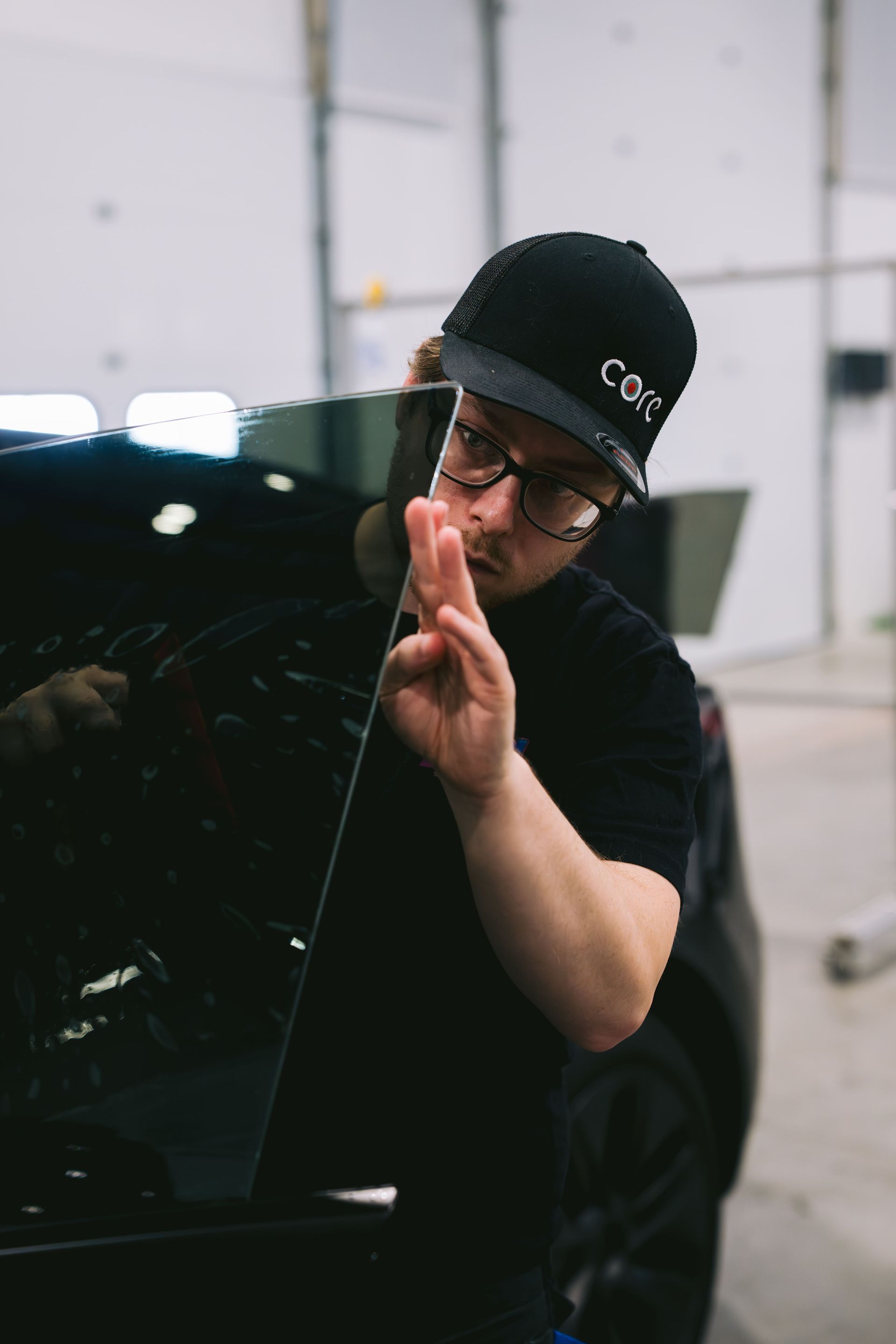 Person in black cap and glasses peeks from behind a car, holding two fingers up in a garage.