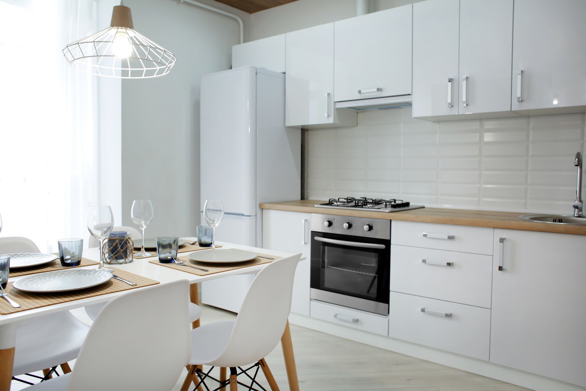 White kitchen with dining table set for a meal. Includes cabinets, appliances, and a hanging light fixture. White kitchen with dining table set for a meal. Includes cabinets, appliances, and a hanging light fixture.