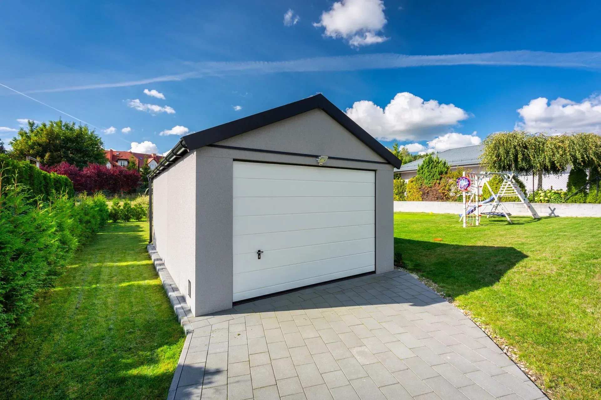 Gray garage with white door on a paved driveway, surrounded by green grass and a blue sky. Gray garage with white door on a paved driveway, surrounded by green grass and a blue sky.
