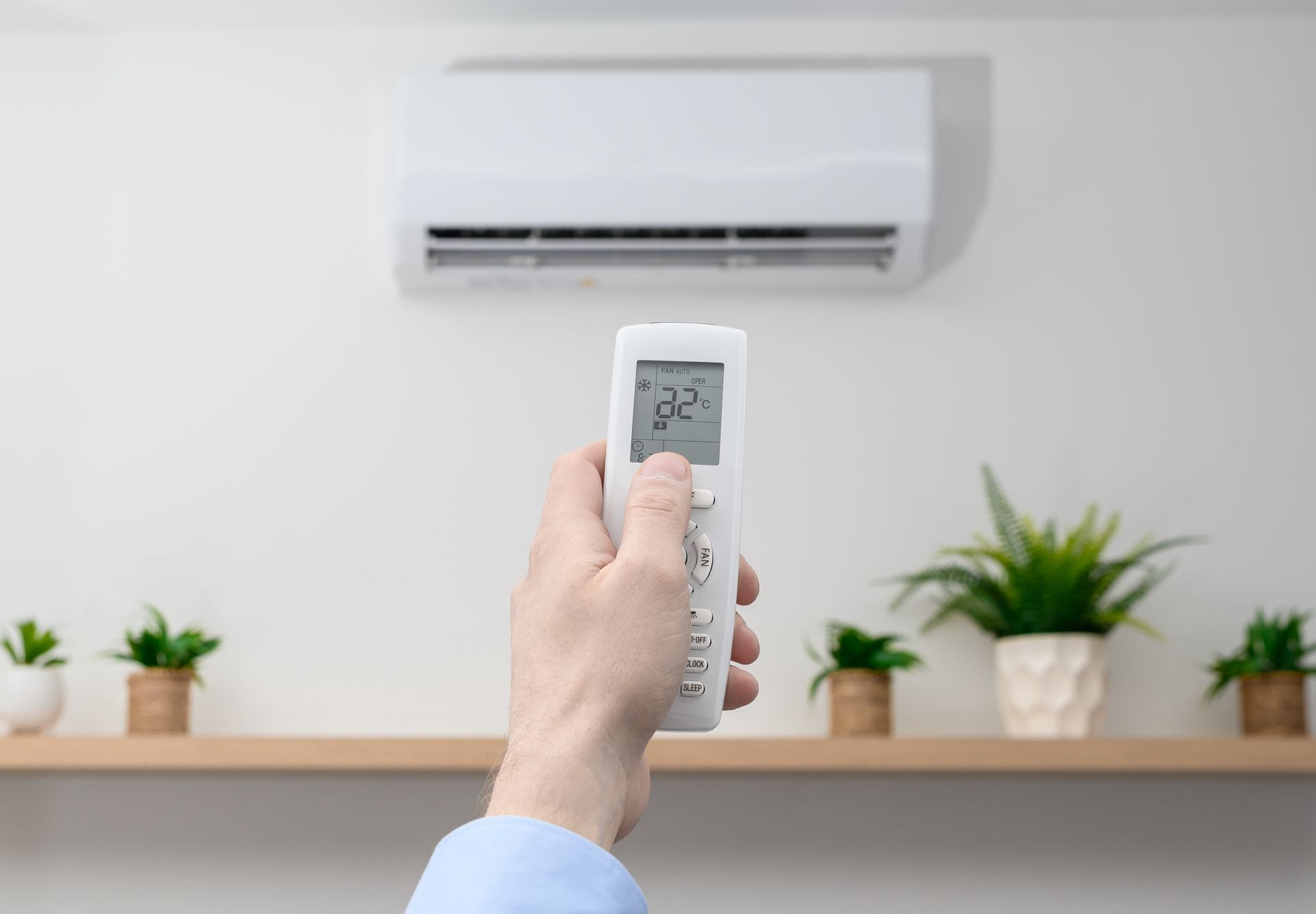 Hand holding a remote aimed at a wall-mounted air conditioner in a bright room