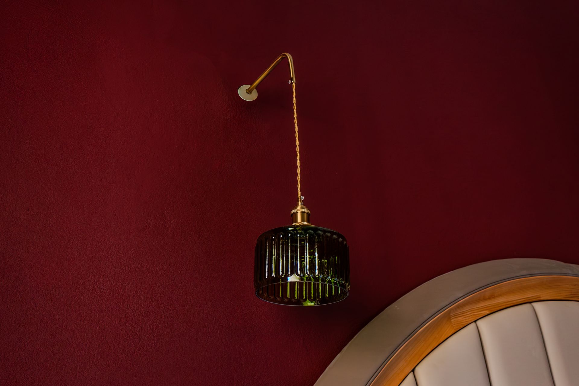 A gold wall lamp with a black cage shade on a deep red wall beside a white upholstered chair