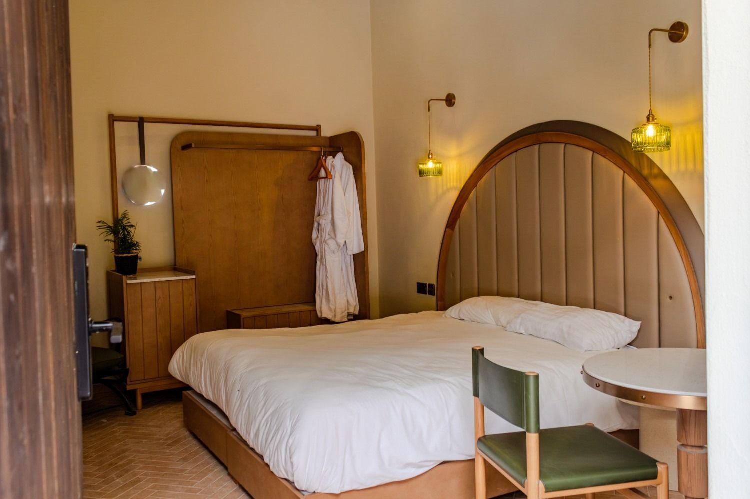 Cozy bedroom with white bed, arched wood headboard, warm wall lights, and a small table and chair.