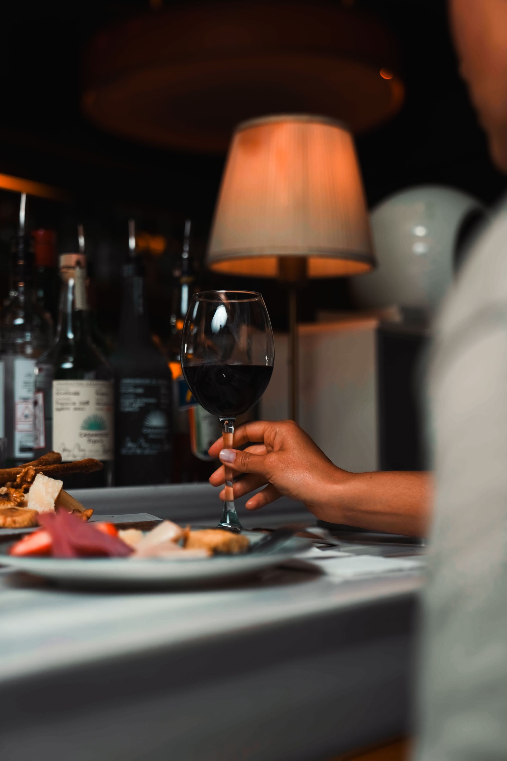 Hand holding a wine glass at a dimly lit dinner table with plates and a glowing lamp