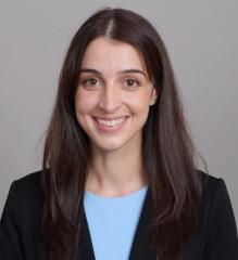 Smiling person in a black blazer and light blue top against a gray background