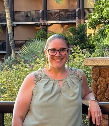 Smiling person in glasses standing outdoors beside tropical plants and a stone wall.