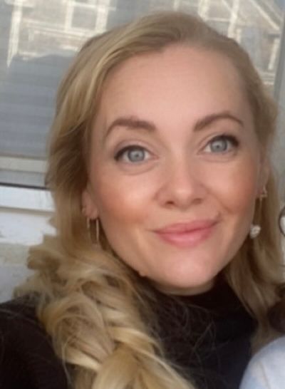 Close-up portrait of a smiling blonde woman in a black top, indoors by a bright window.