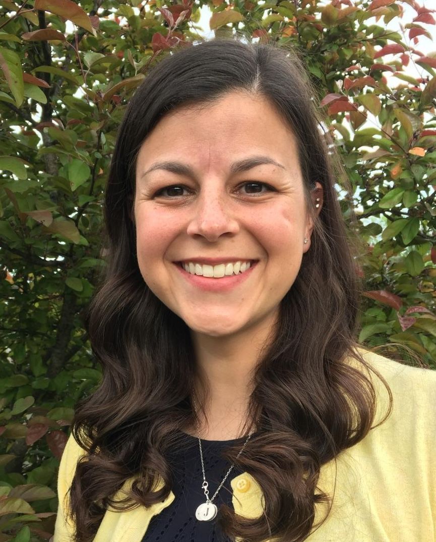Smiling woman with dark hair standing outdoors in front of leafy trees, wearing a yellow cardigan and necklace