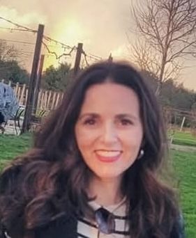 Smiling woman with dark hair outdoors at sunset, wearing a striped top in a grassy park.