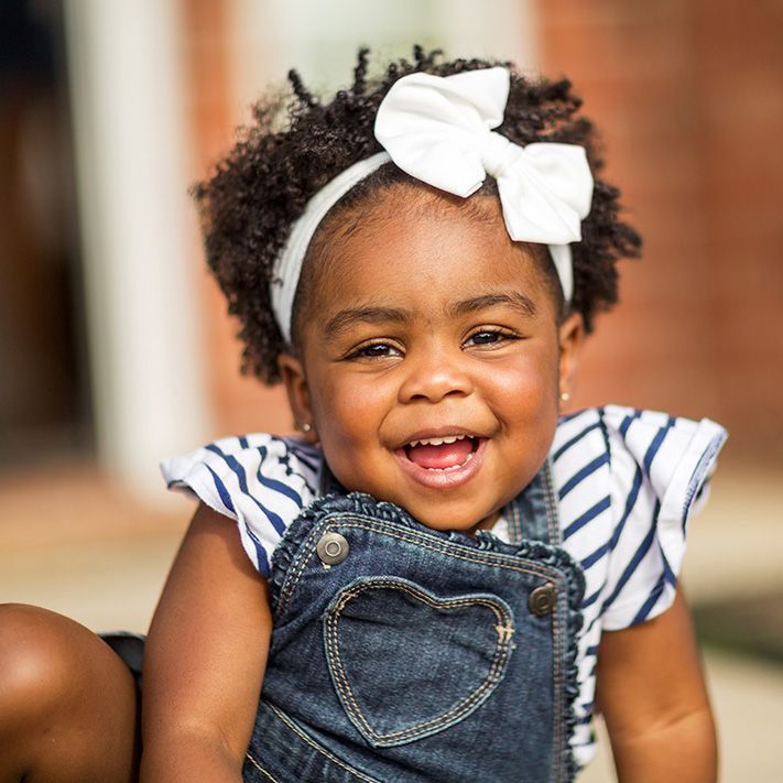Smiling child with white bow and striped shirt under denim overalls indoors