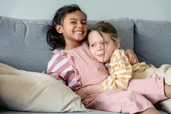 Two children sitting on a couch, smiling and cuddling together in soft pink and striped pajamas.