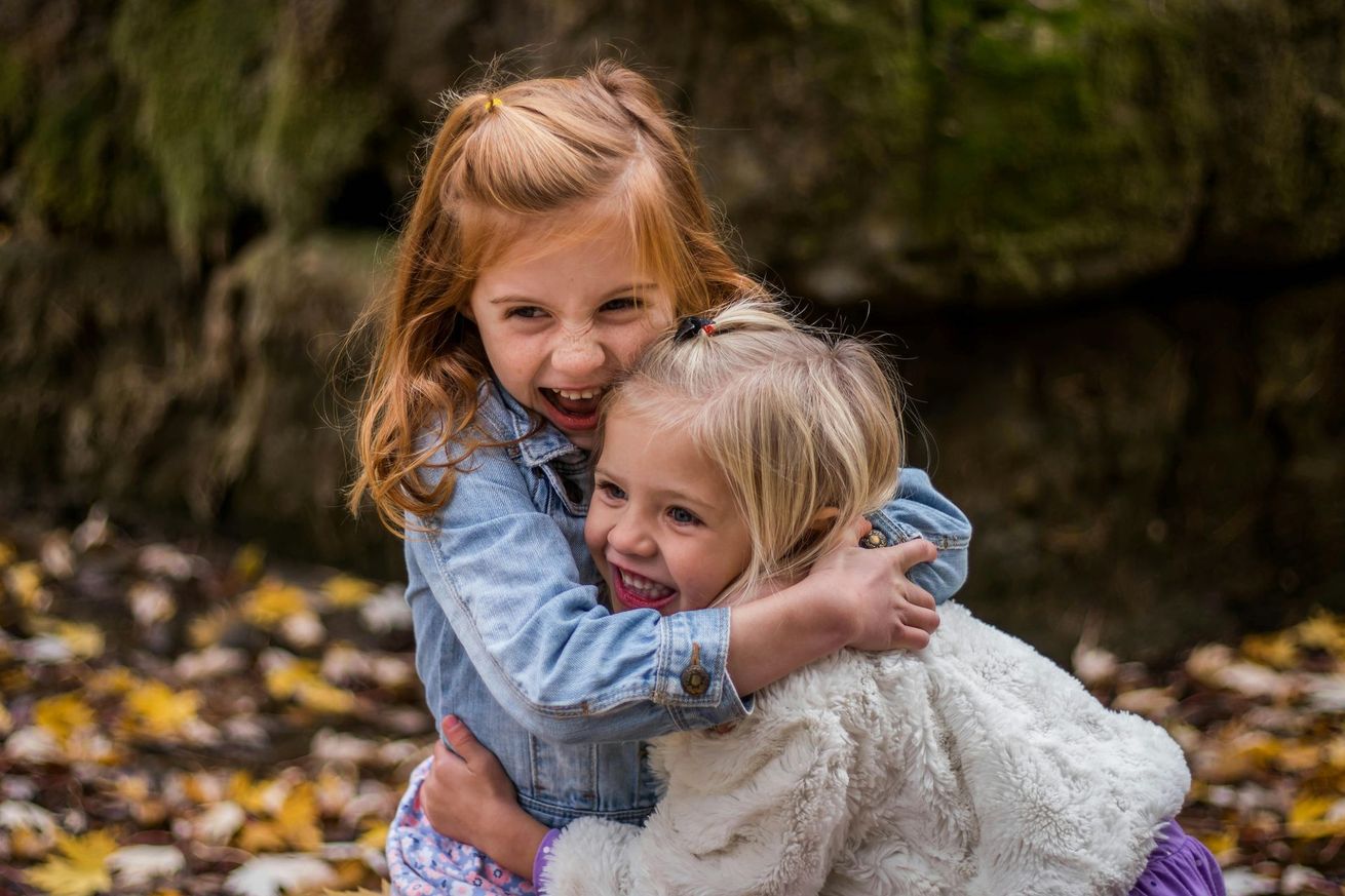 Two children hugging and smiling outdoors, one in a denim jacket and the other in a white sweater.