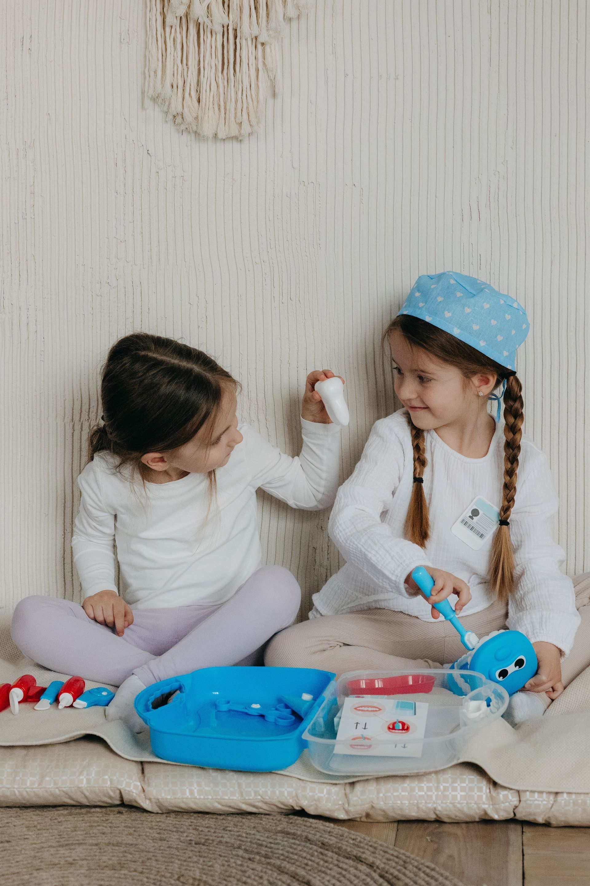 Two children in white clothes play with toys on a cushioned floor, smiling at each other.