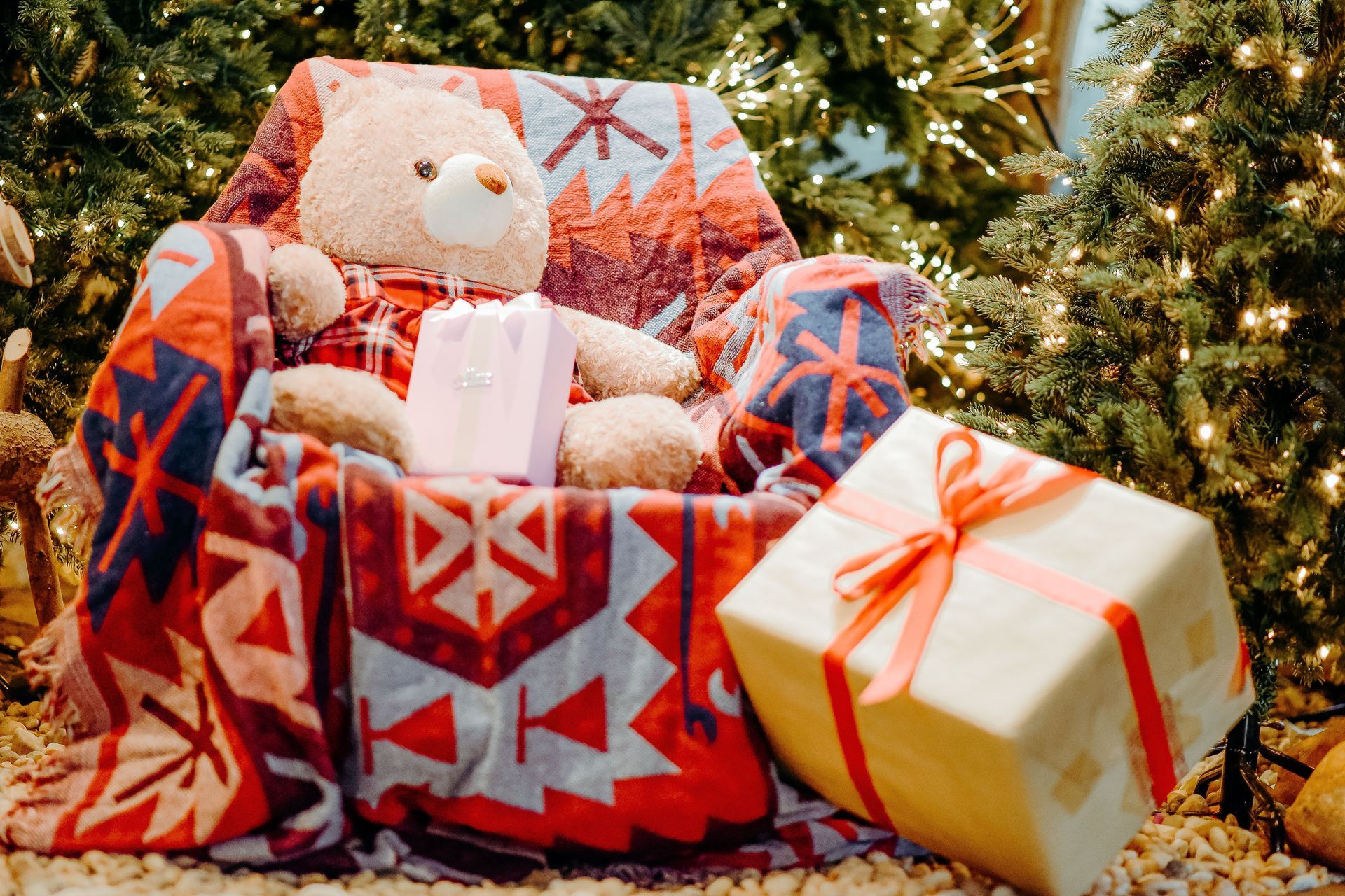 Teddy bear in a red patterned chair beside wrapped gifts and a holiday tree