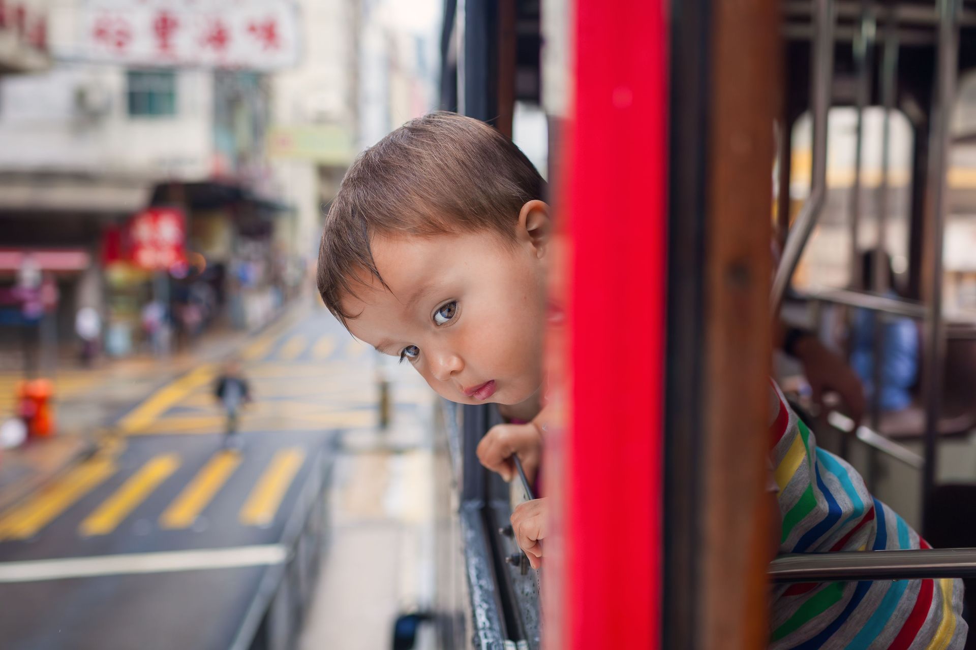 Child leaning out of a red vehicle, looking curiously at a blurred city street.