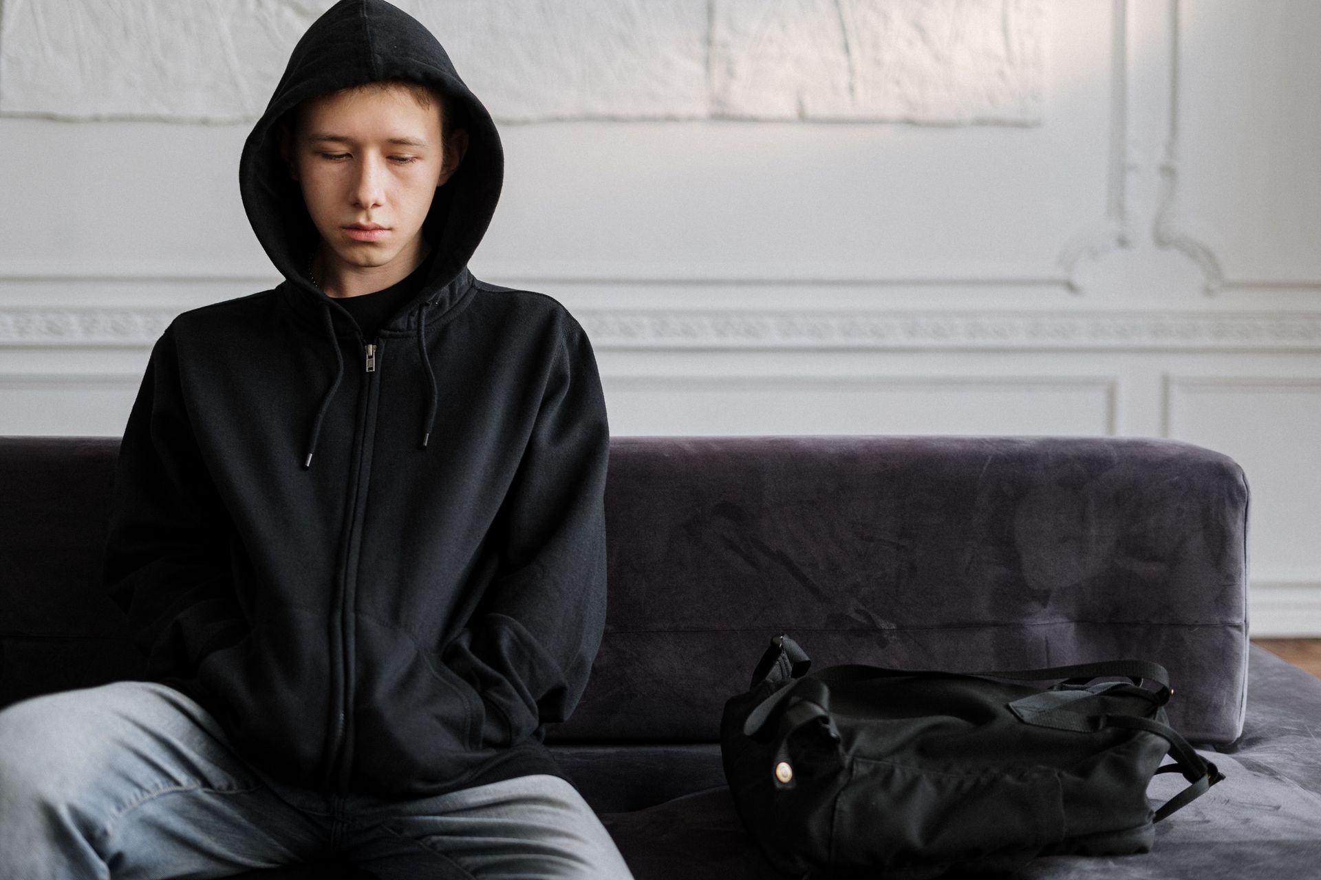 Person in a black hoodie sits on a gray couch beside a black bag in a bright room.