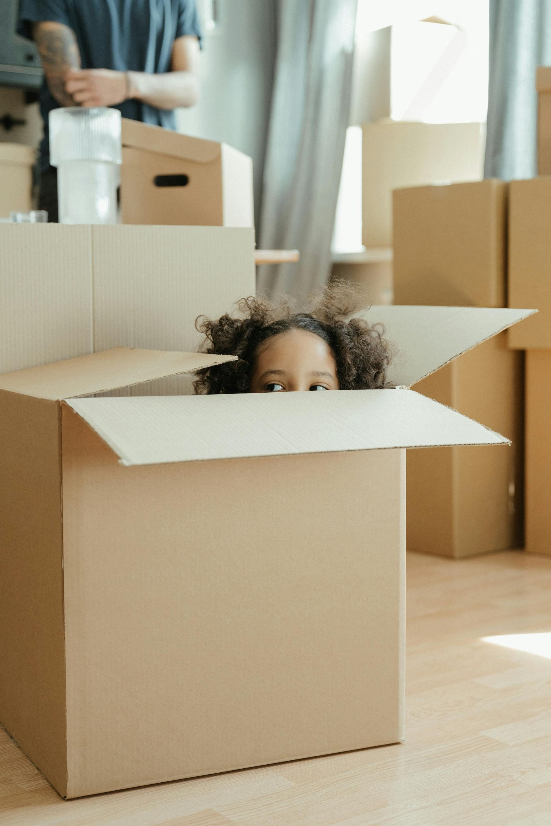 Child peeking out of a cardboard box in a bright room with moving boxes and curtains
