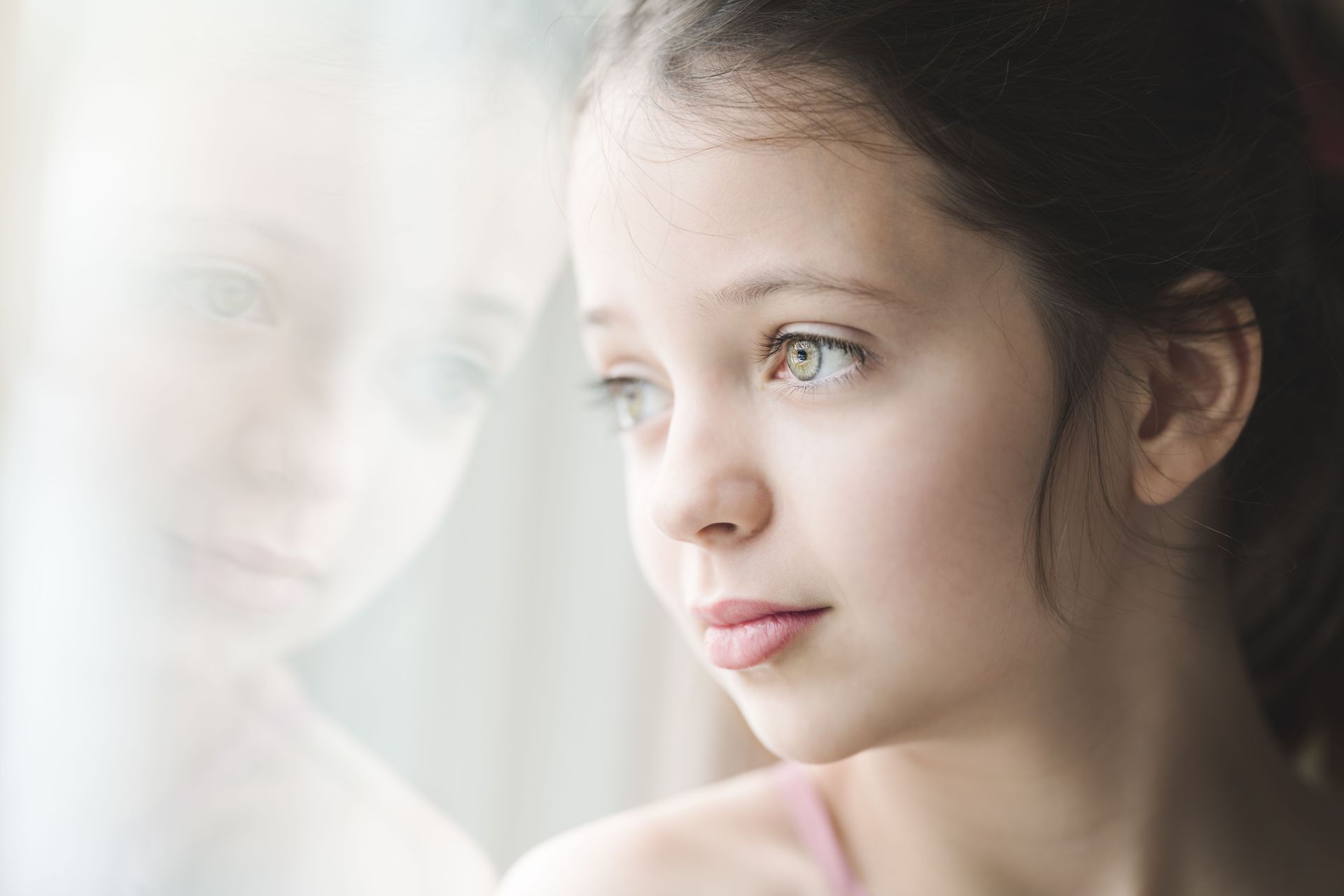 A young person gazes out a window, her reflection faintly visible in the glass.