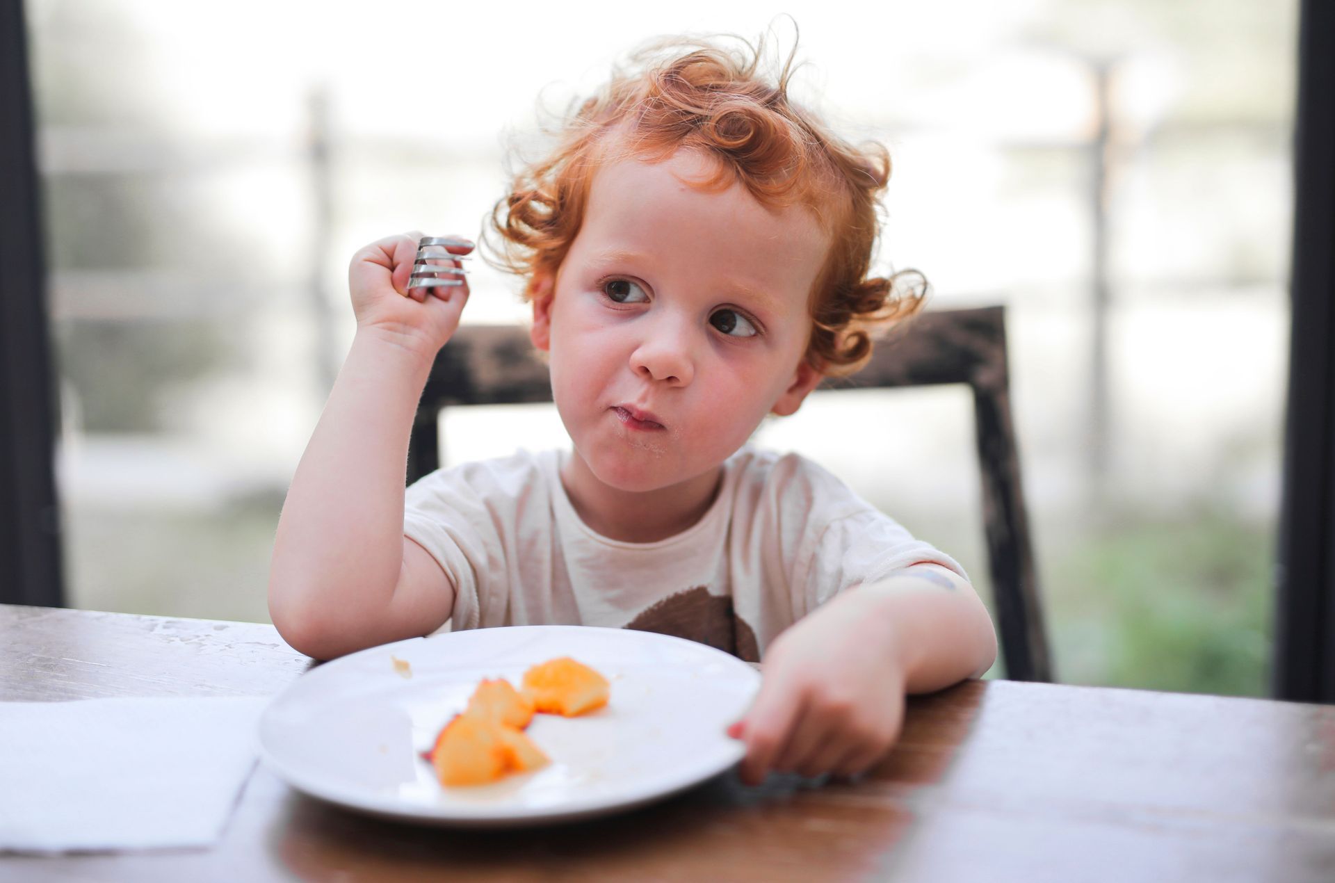 Young child sitting at a table, holding a snack and looking thoughtfully at a plate of orange pieces.