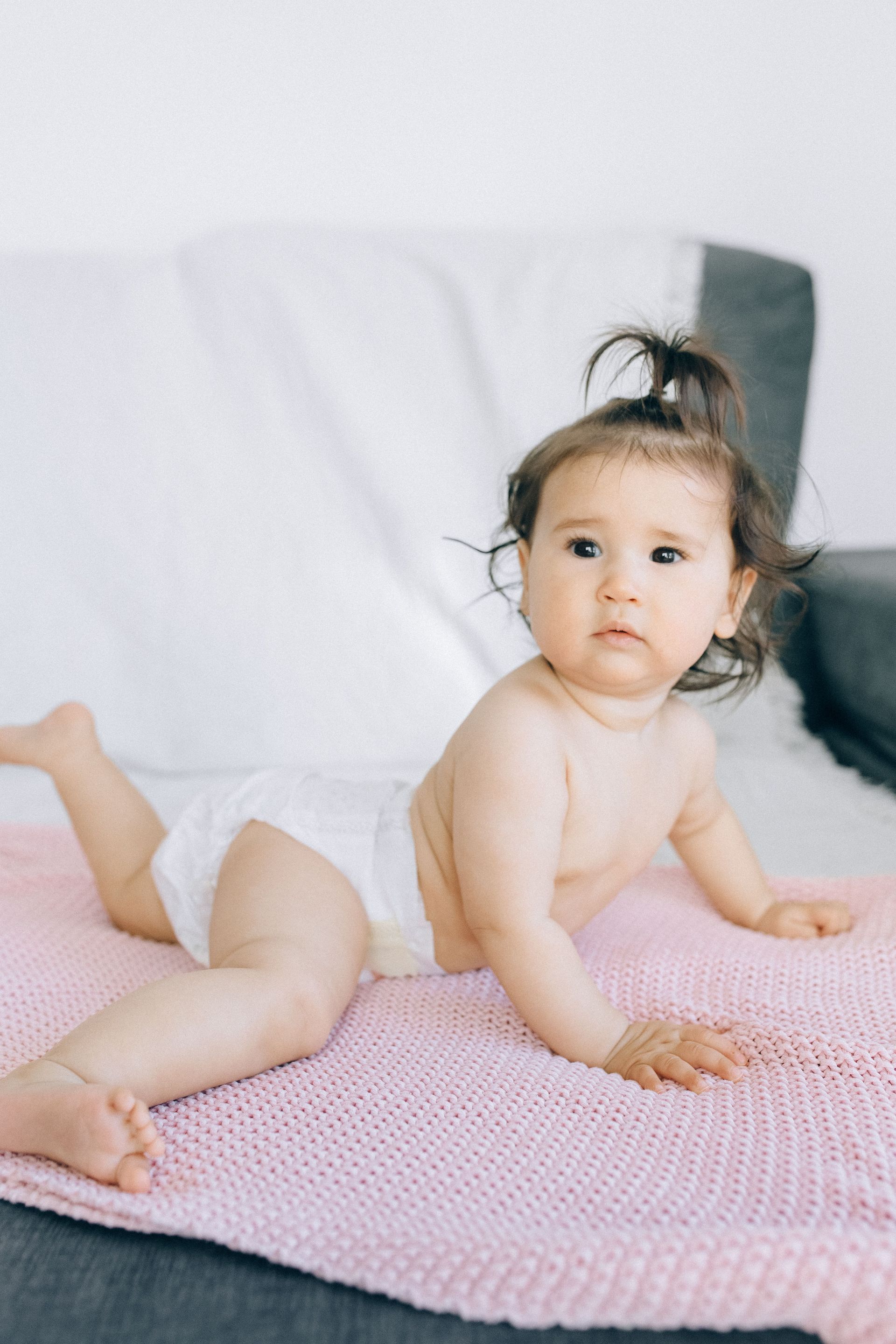 Toddler on a pink blanket, propped on hands and looking at the camera in a bright room
