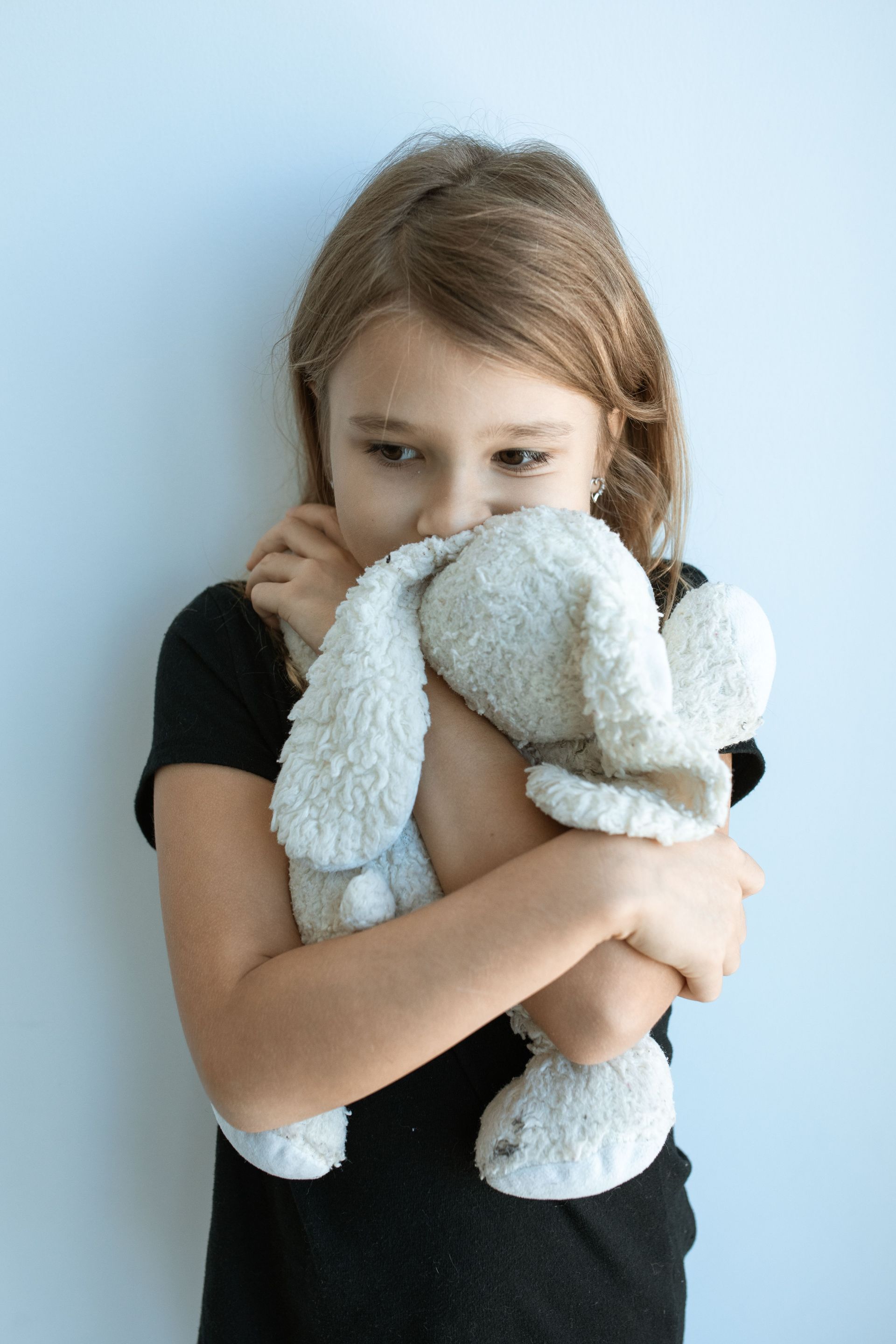 Child in a black shirt hugging a white stuffed rabbit against a light blue background