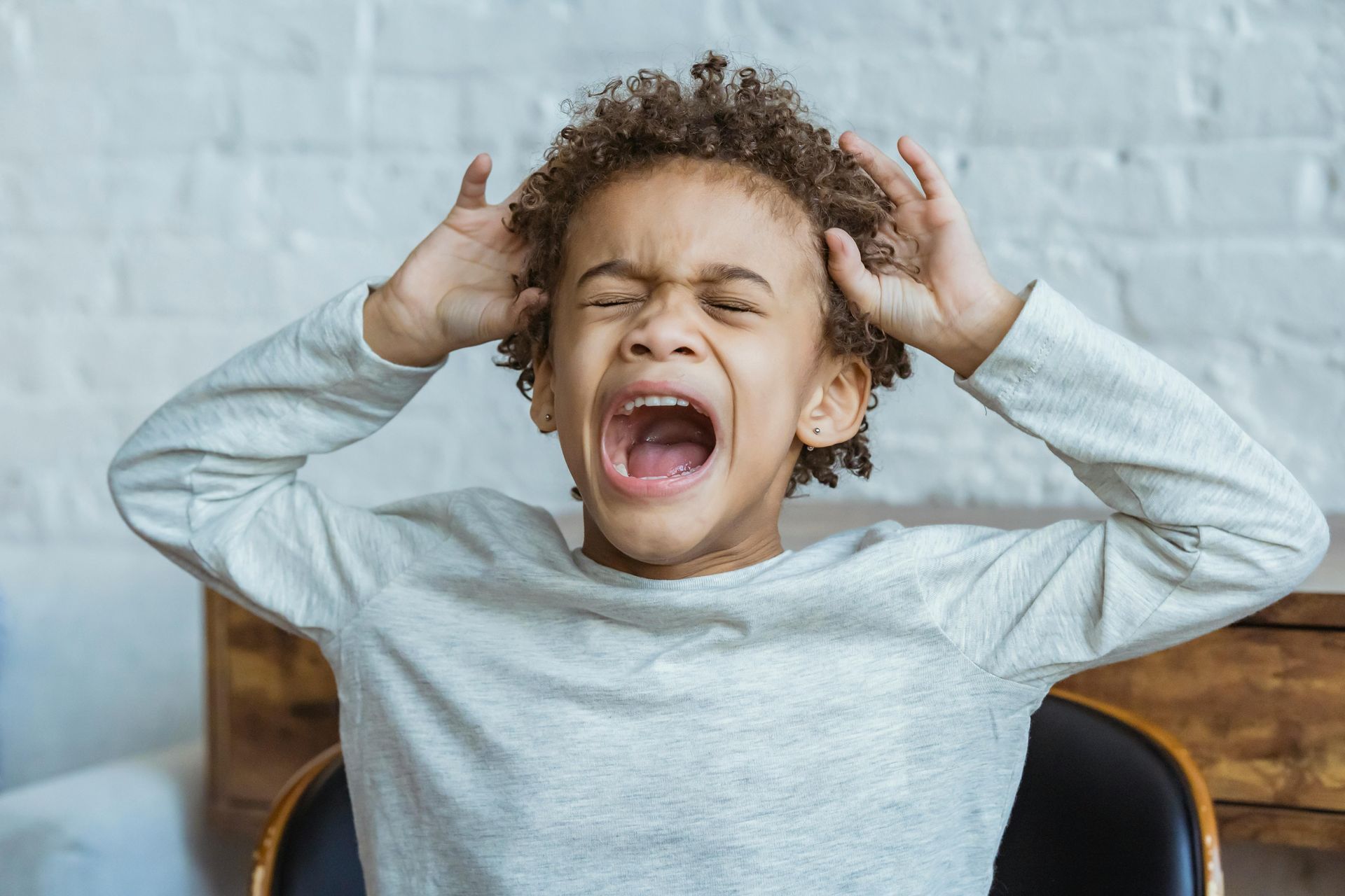 Person yawning with hands by ears, eyes closed, wearing a light gray sweater on a chair.