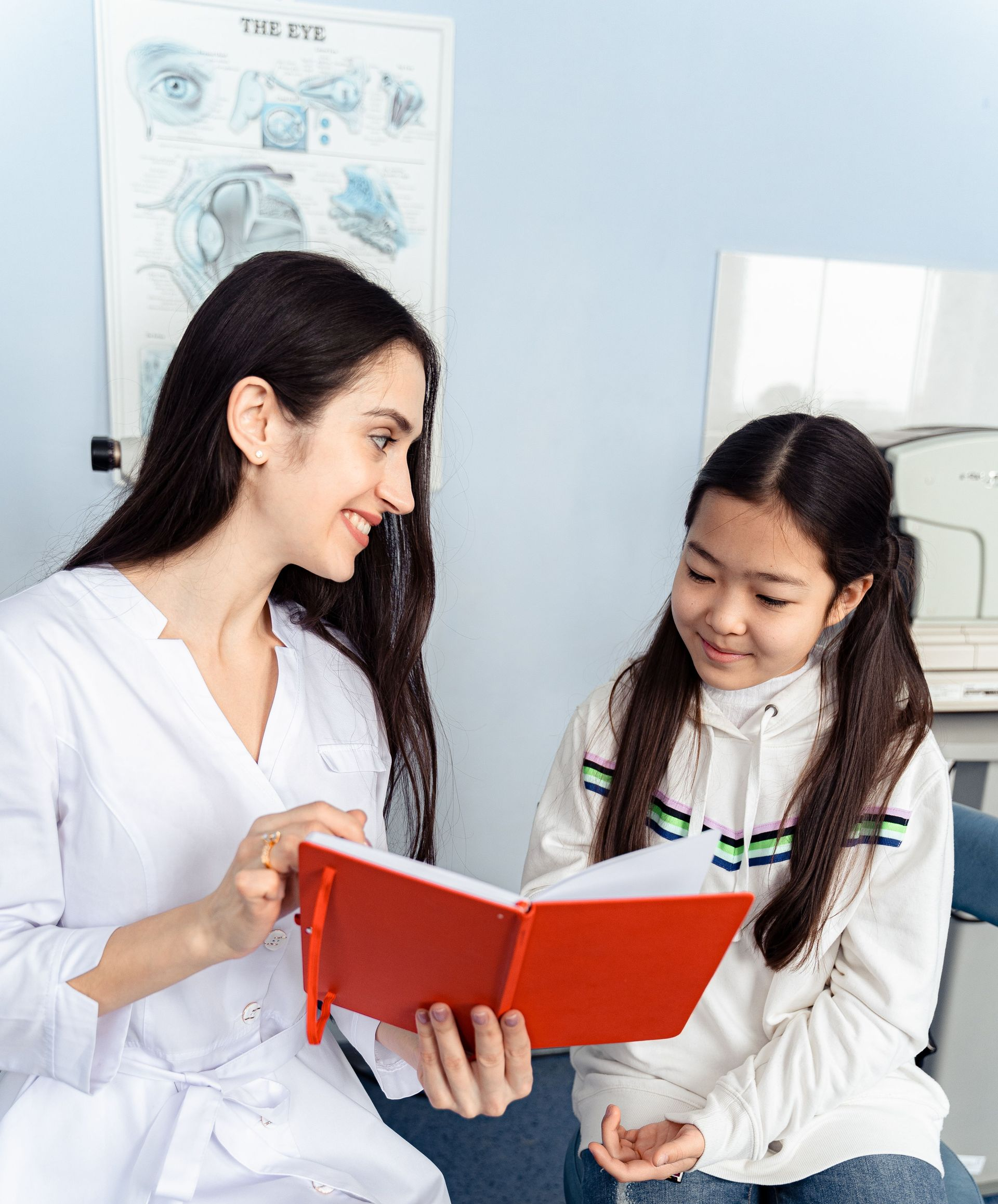 Two people in white coats smiling and reading a red book together in a bright room.