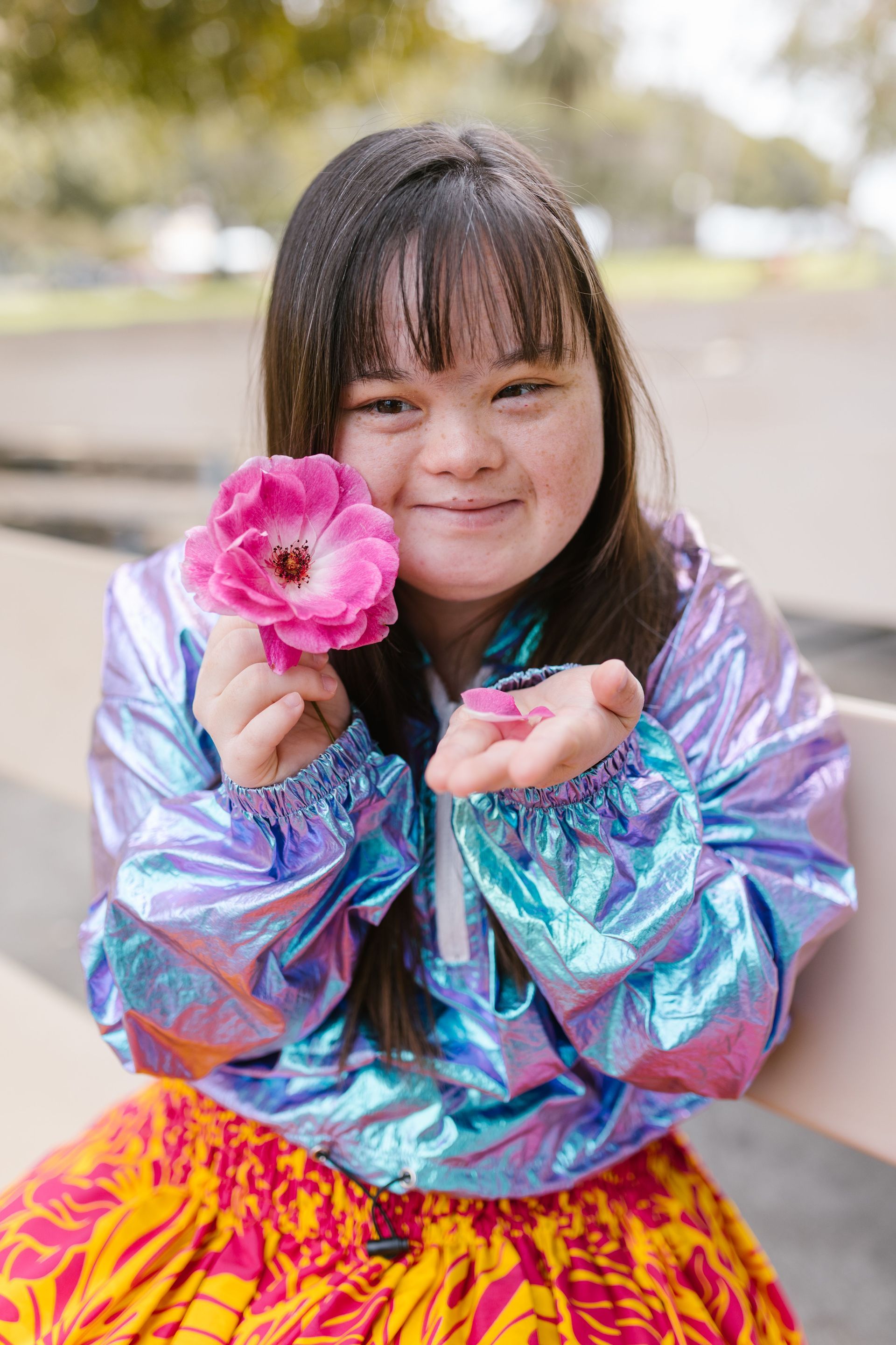 Smiling child holding a pink flower, wearing a shiny iridescent jacket and bright patterned skirt outdoors