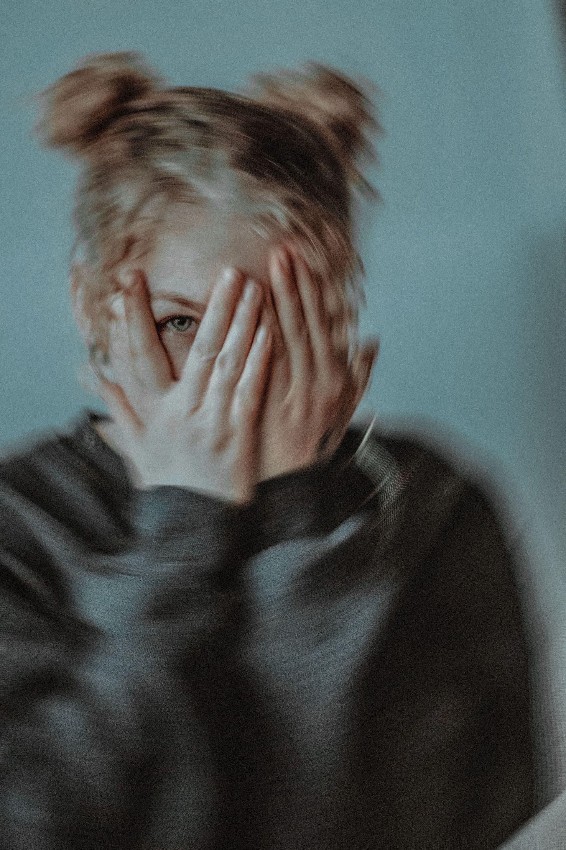 Person with messy hair covering their face with both hands against a muted blue background.