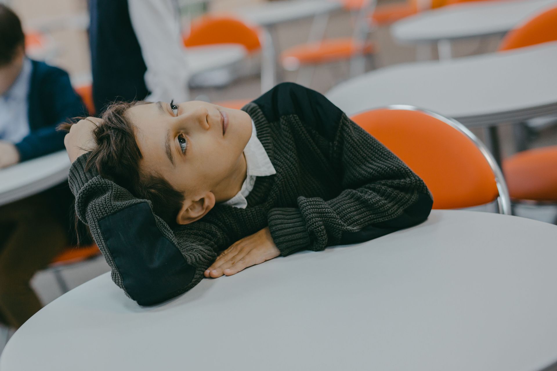 Child leaning on a round table, looking up in a classroom with orange chairs.