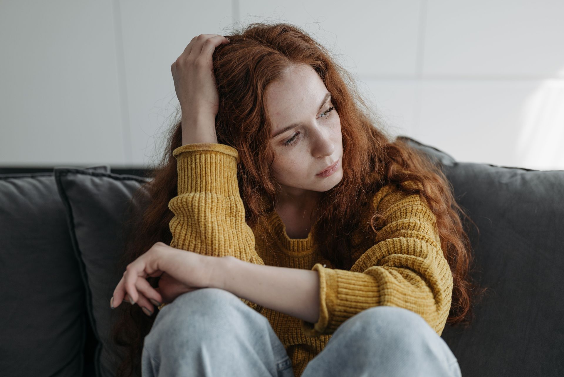 Woman with red hair sitting on a couch, leaning her head on her hand and looking down thoughtfully.