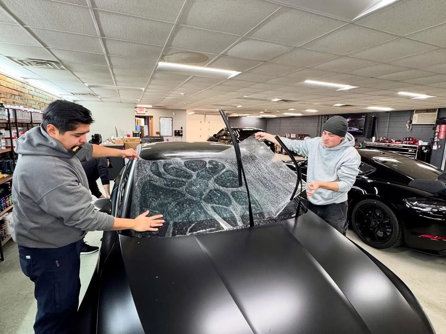 Two workers in a garage install clear protective film on a black car hood and windshield.