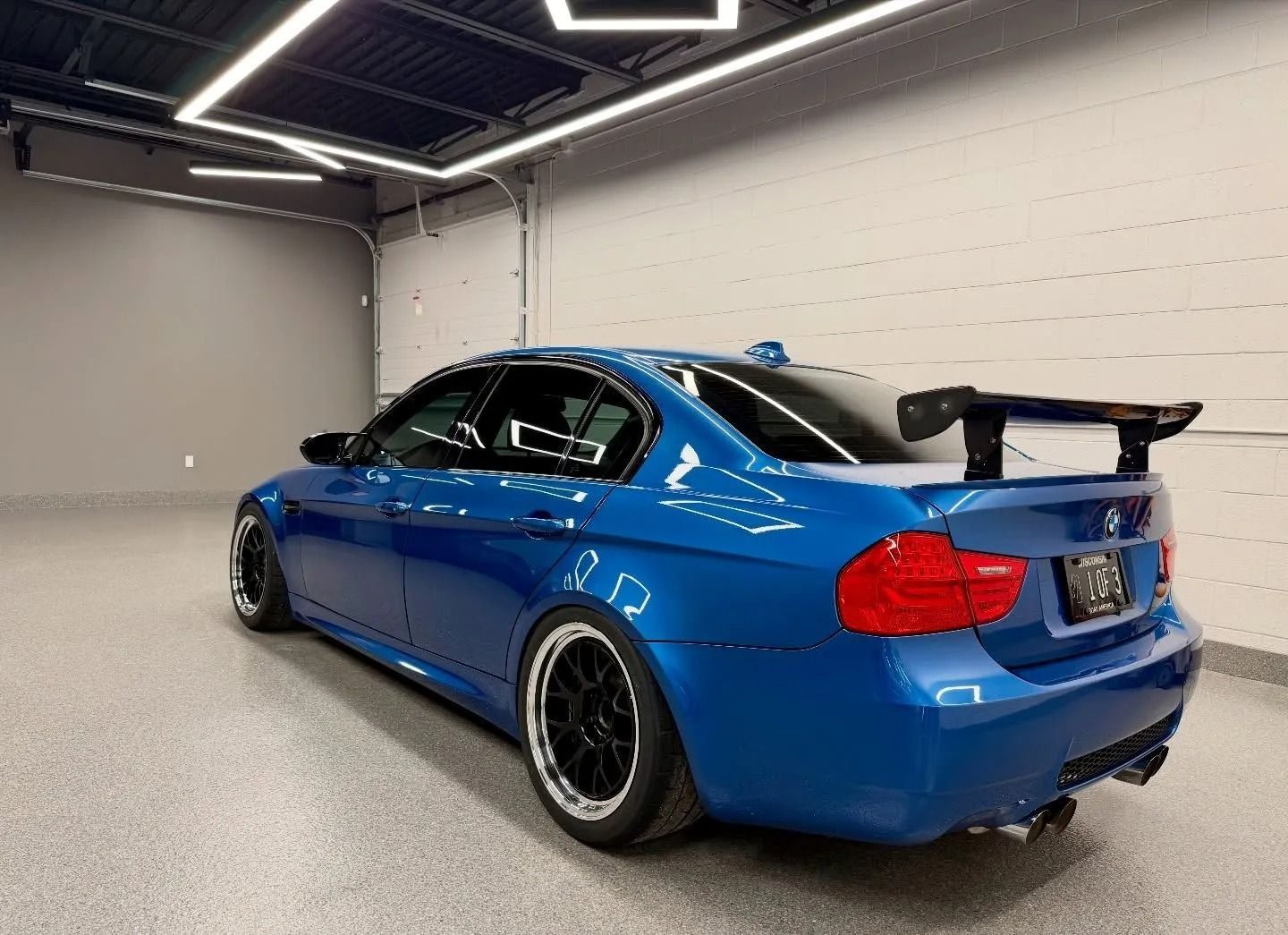 Blue sports coupe with a large rear wing in a garage-like showroom, rear three-quarter view.