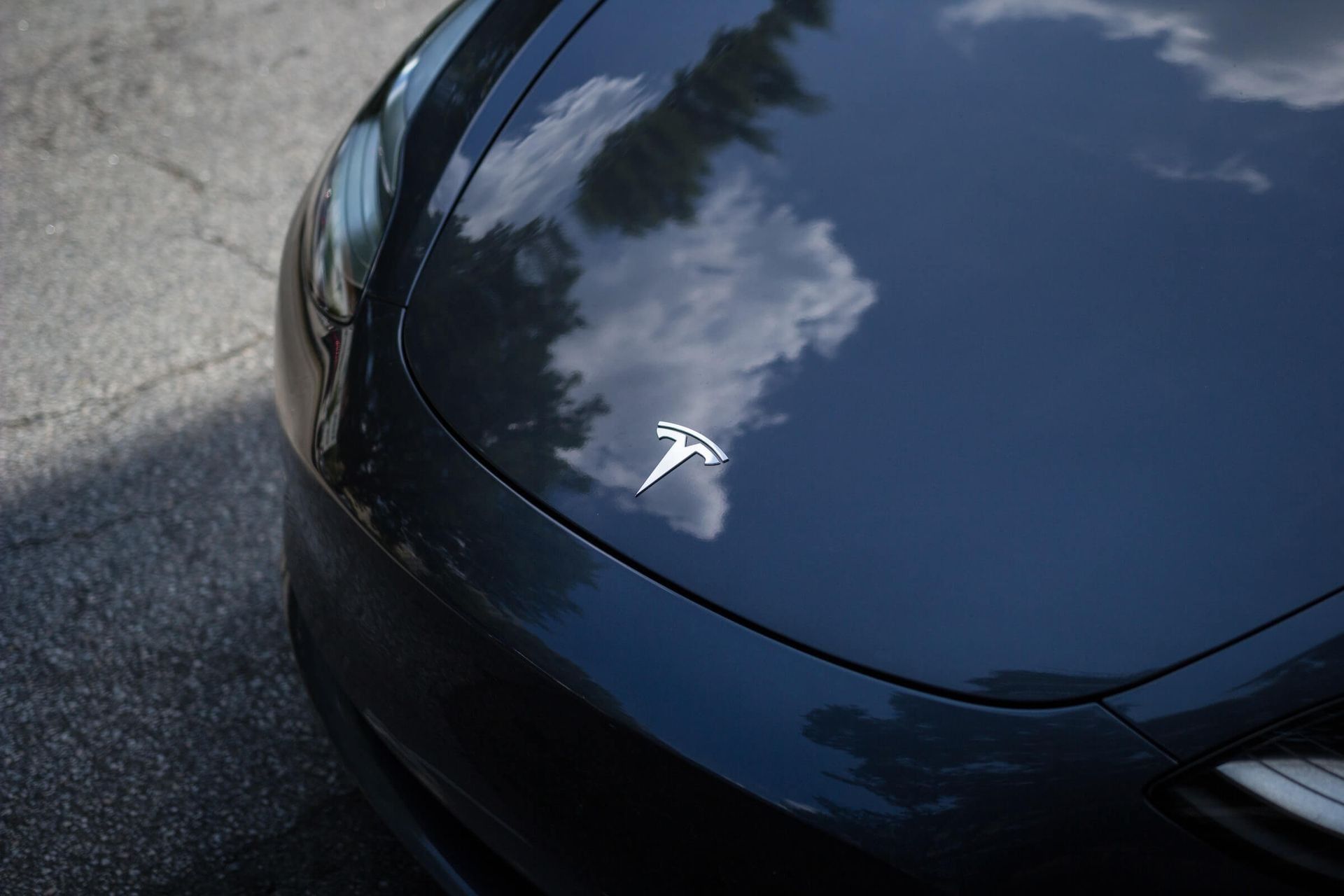 Close-up of a black Tesla hood with the silver logo reflecting clouds and sky