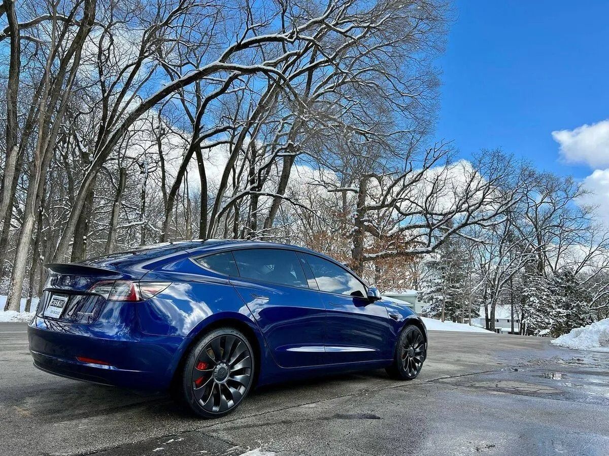 Blue Tesla parked on a snowy driveway beside bare trees under a bright blue sky