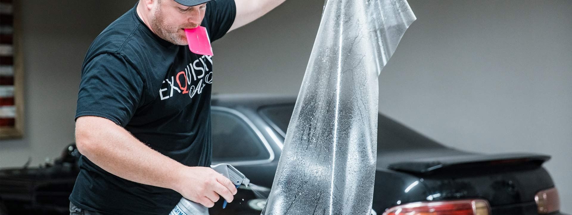 Person washing a black car in a garage, spraying water from a hose while holding a pink cloth.