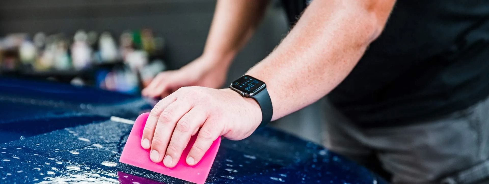 Person wipes a blue car with a pink sponge, close-up of hands on the hood.